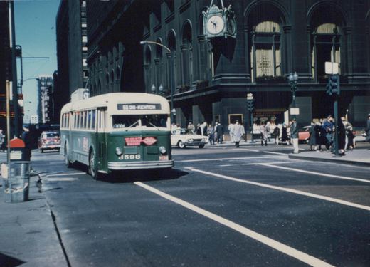 A green and cream Chicago Transit Authority bus, number 3593, is turning at a downtown intersection with pedestrians and classic cars nearby, under a clear blue sky.