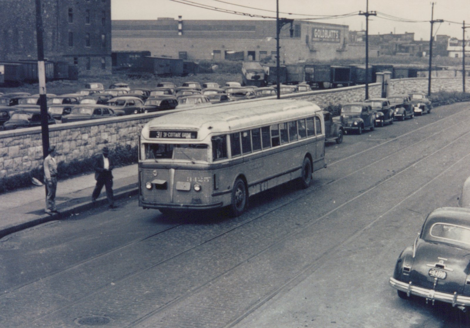 A black and white photo of a vintage motor bus on a city street with trolley tracks, surrounded by 1940s cars and two pedestrians on the sidewalk.