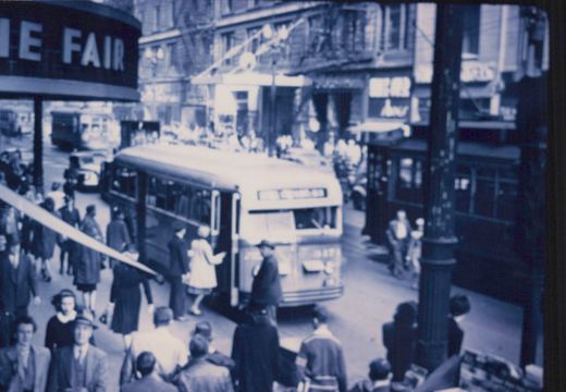 A vintage bus with the destination 'MILWAUKEE 63' and fleet number 2874 is surrounded by pedestrians on a city street, with storefronts and a street sign visible in the background.