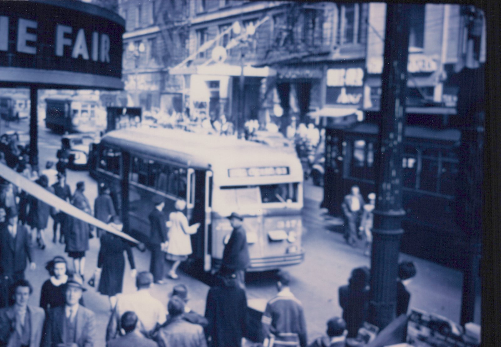 A vintage bus with the destination 'MILWAUKEE 63' and fleet number 2874 is surrounded by pedestrians on a city street, with storefronts and a street sign visible in the background.