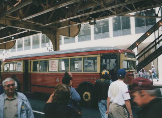 A vintage red and cream bus with 'Illinois Railway Museum' on the side is parked under an elevated railway, with several people gathered around it.