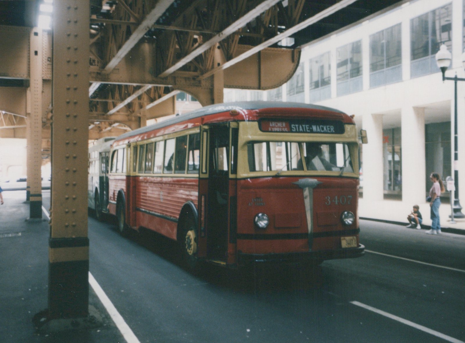A red and cream vintage bus, number 3407, is parked under a metal elevated train structure on a city street, with a few people on the sidewalk and another bus behind it.
