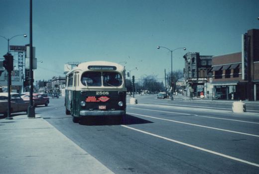 A vintage city bus, number 2508, is shown driving down a city street with storefronts and parked cars on either side under a clear sky.