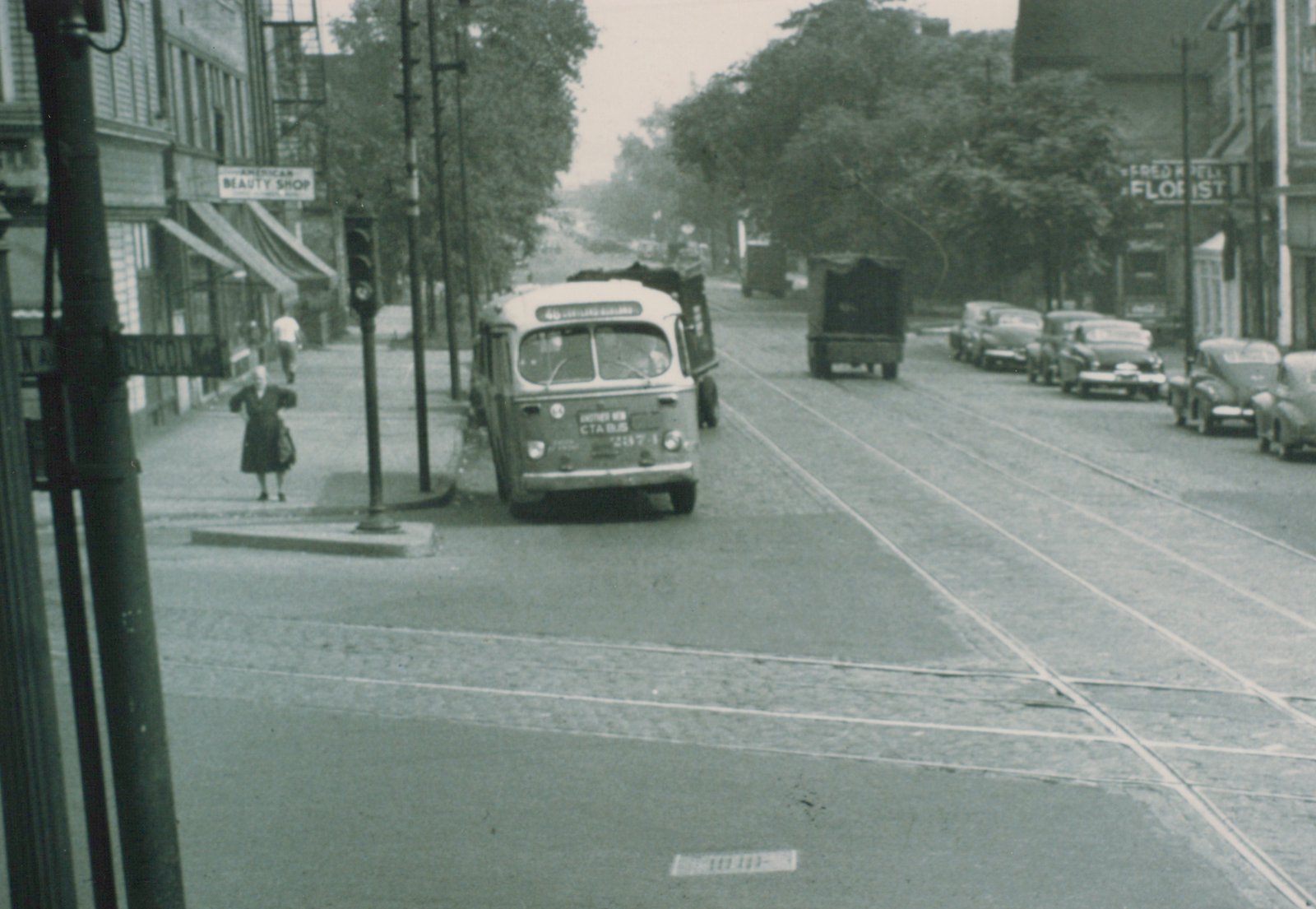A vintage black-and-white photo of a CTA bus driving down a street with trolley tracks, past storefronts and parked 1950s cars.