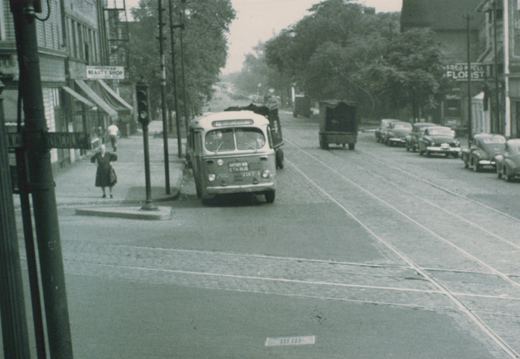 A vintage black-and-white photo of a CTA bus driving down a street with trolley tracks, past storefronts and parked 1950s cars.