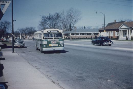 A green and cream city bus with the number 1806 and route 84 on its sign drives down a paved street on a sunny winter day, with parked and moving cars and bare trees lining the road.