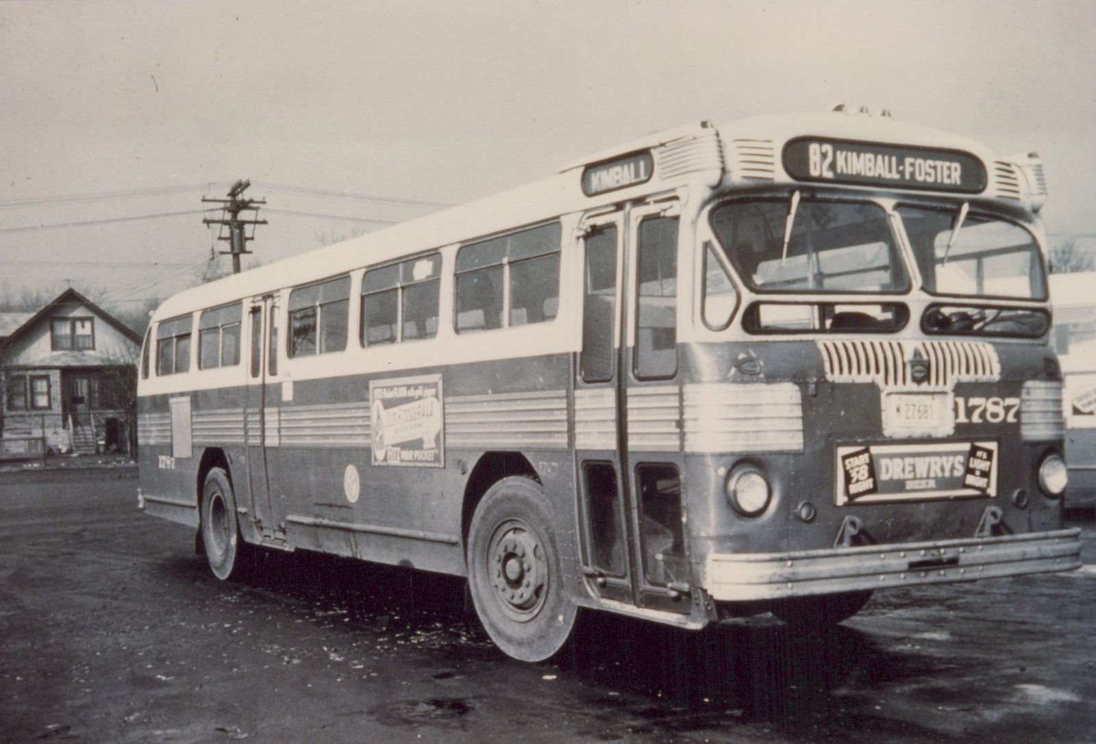 A black and white photo of a Chicago Transit Authority motor bus, number 1787, showing its front and side with route signs and advertisements, parked on a wet street with a house in the background.