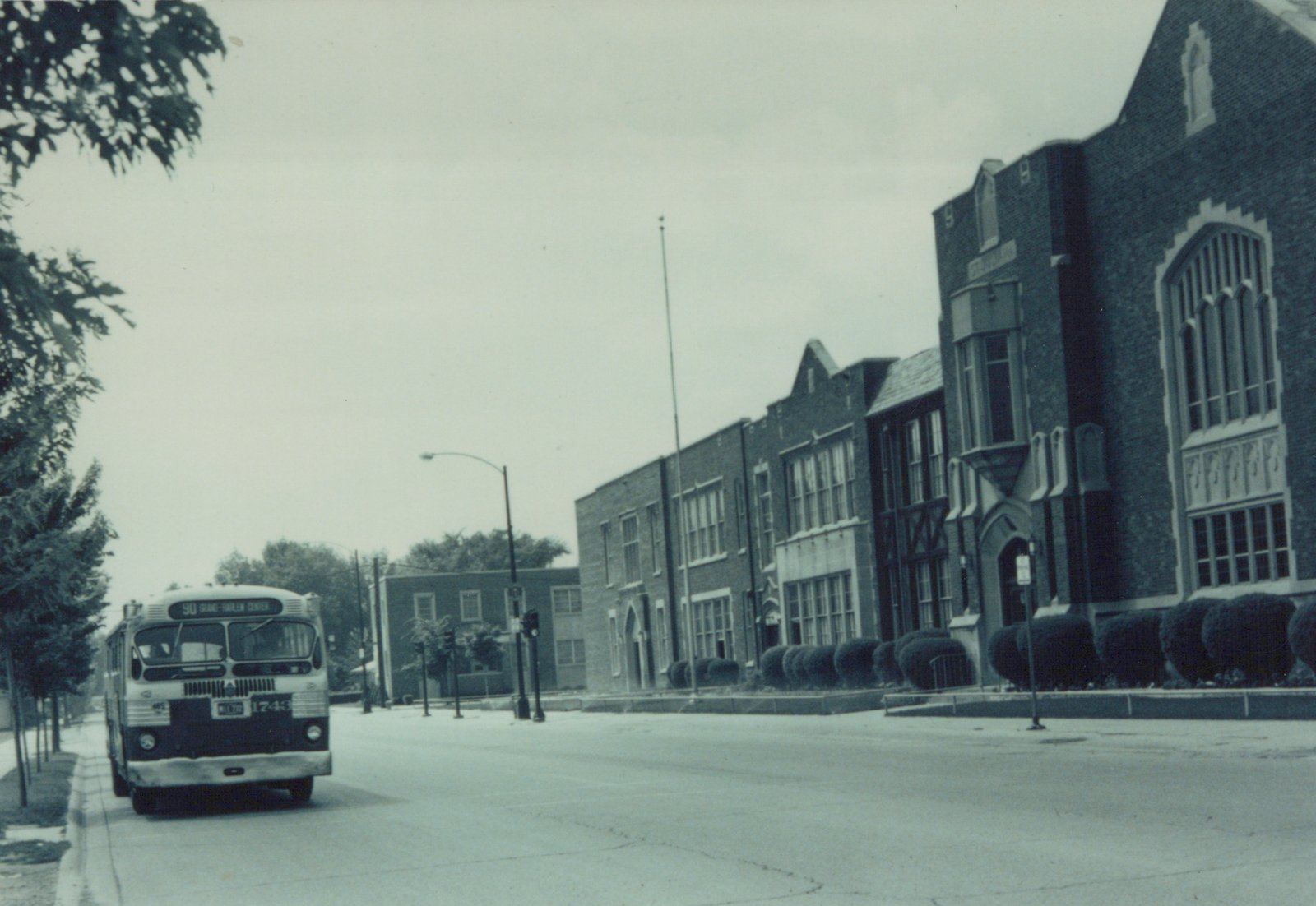 Front view of a vintage white and dark-colored bus with route 90 Grand-Harlem Center on its sign, driving on a city street next to a large brick building under an overcast sky.