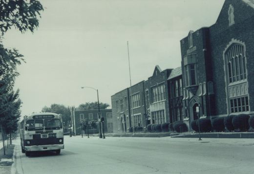 Front view of a vintage white and dark-colored bus with route 90 Grand-Harlem Center on its sign, driving on a city street next to a large brick building under an overcast sky.