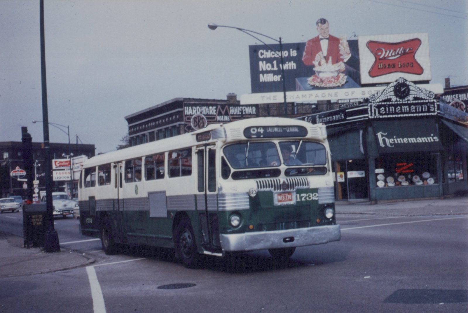 A green and cream Chicago Transit Authority bus with the number 1732 is turning a corner on a city street, with storefronts and a large billboard for Miller beer in the background.
