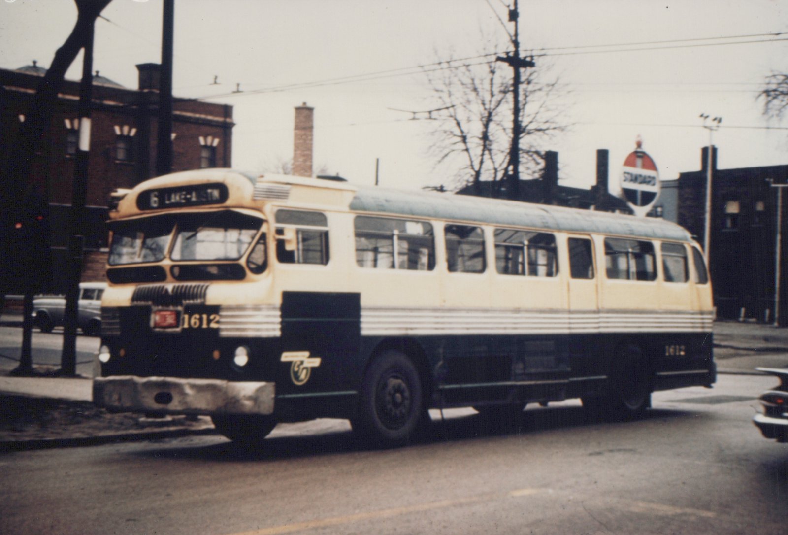 A cream and dark green CTA bus with the number 1612 on its front and rear, showing the destination '6 LAKE-AUSTIN' on its sign, driving down a street with a Standard Oil sign and bare trees in the background.