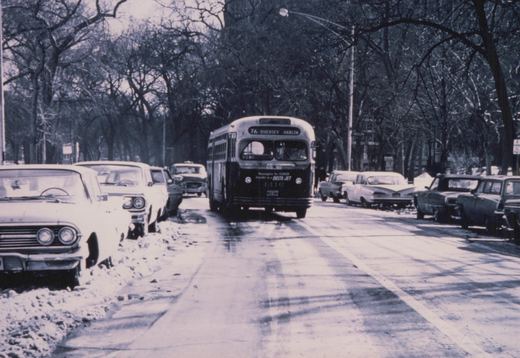 A black and white photo of a 1960s-era CTA bus driving down a snow-covered street lined with parked cars and bare trees.