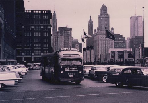 A black and white photo of a Chicago Transit Authority bus, number 1353, with a 'Shuttle Service' sign, driving down a city street lined with 1950s cars and tall buildings in the background.