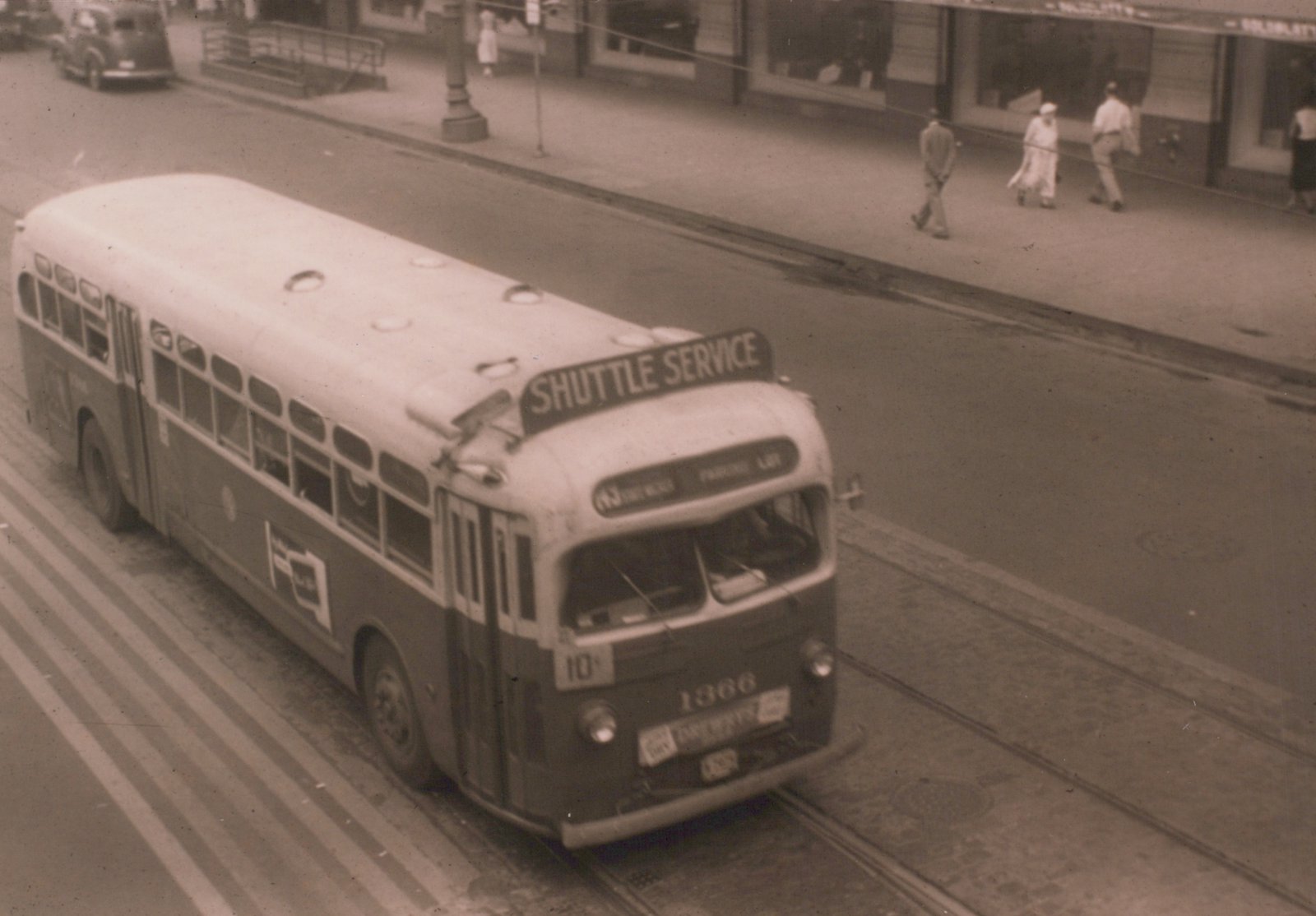A vintage motor bus with the number 1366 and 'SHUTTLE SERVICE' on its roof sign, traveling on a city street with visible trolley tracks.
