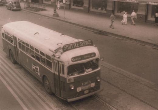 A vintage motor bus with the number 1366 and 'SHUTTLE SERVICE' on its roof sign, traveling on a city street with visible trolley tracks.