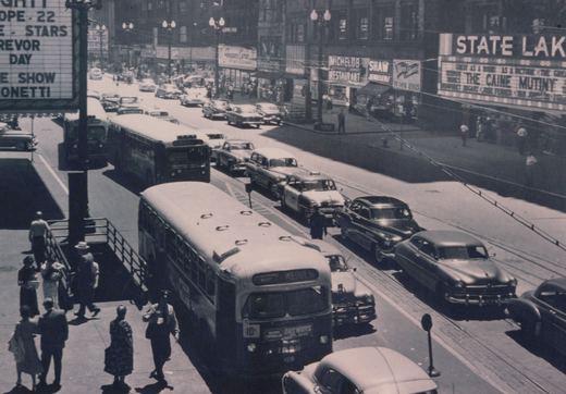 A black and white photo of a Chicago Transit Authority bus from the 1950s on a crowded city street, surrounded by pedestrians, vintage cars, and theater marquees.