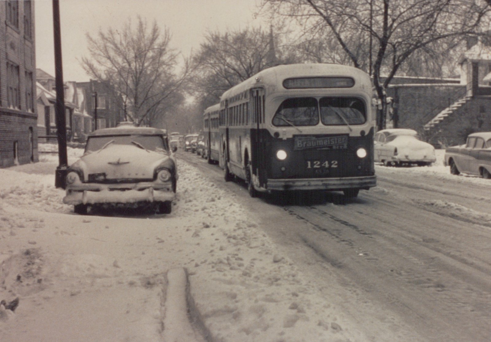 A black and white photo of a mid-1950s Chicago bus driving down a snowy street lined with parked cars and bare trees.