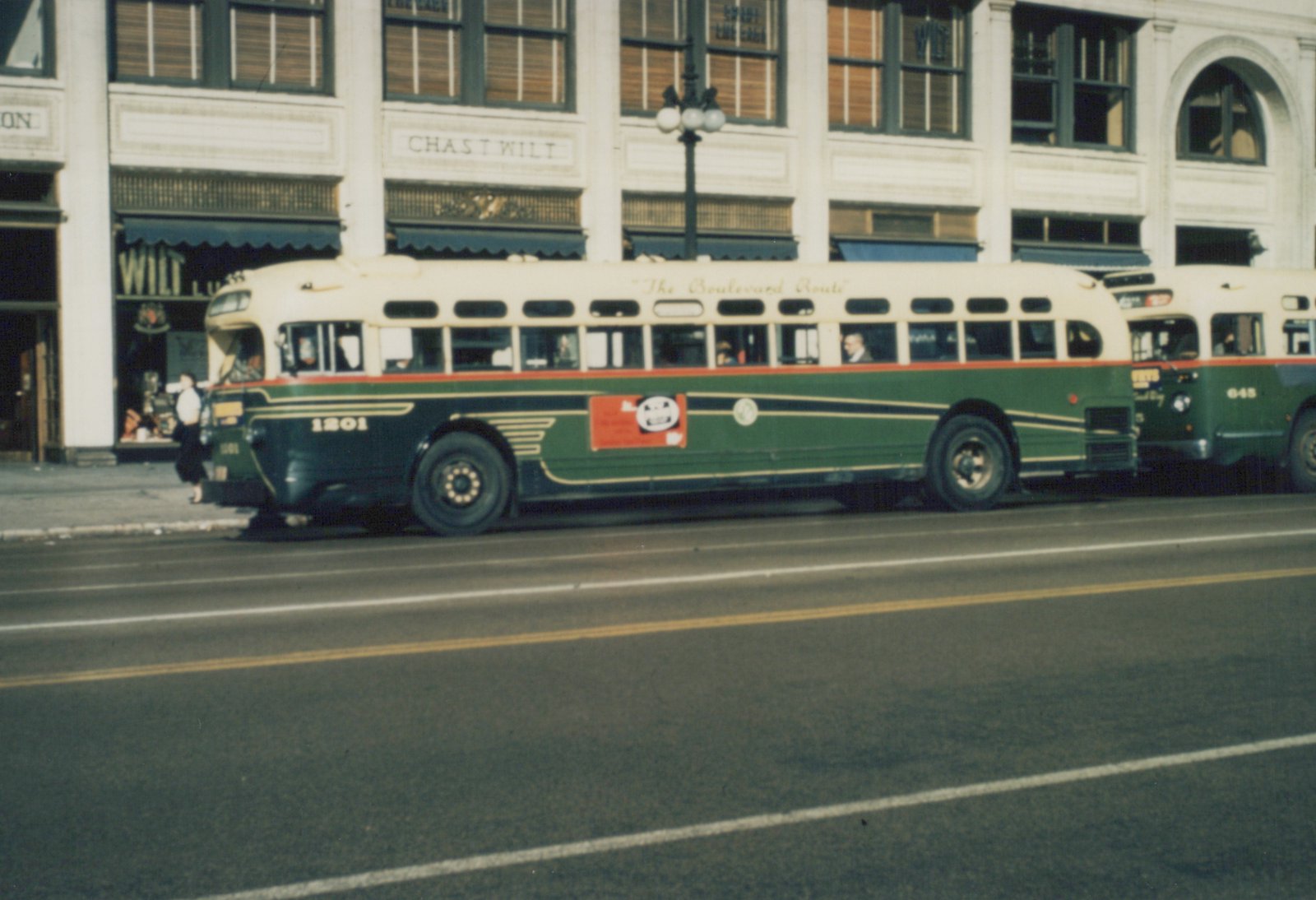 Side view of two green and cream Chicago Surface Lines buses, numbered 1201 and 645, on a city street with a building labeled CHAS T WILT behind them.