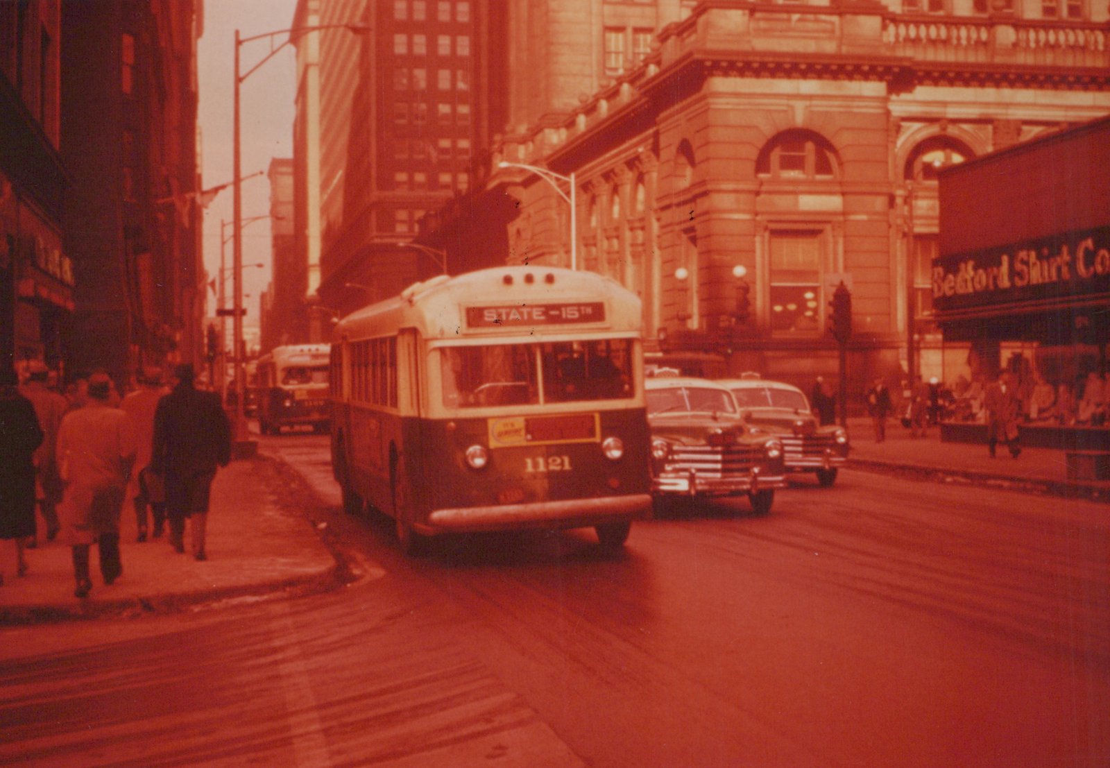A vintage CTA bus numbered 1121 is shown on a city street with pedestrians and classic cars, in front of a building with a sign for Bedford Shirt Co.