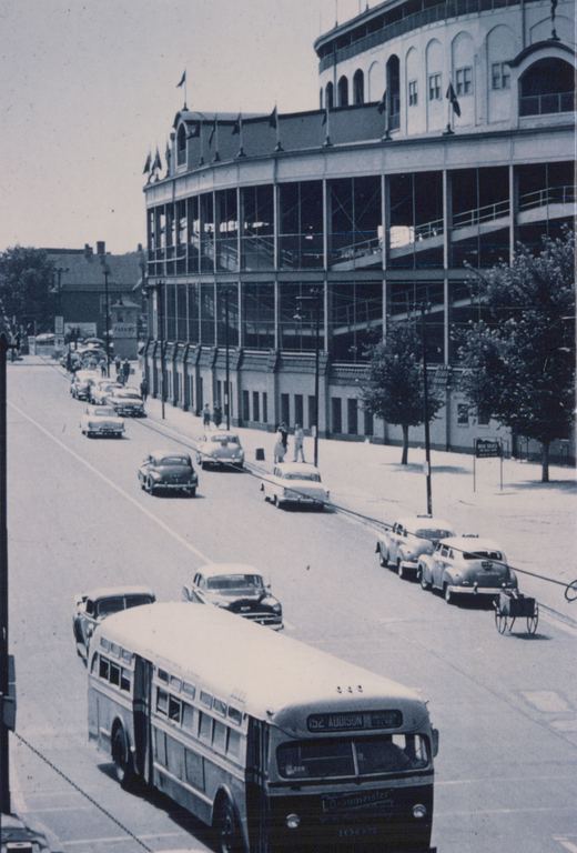 A black and white photo from the 1950s shows a CTA bus on a city street with Wrigley Field in the background, surrounded by classic cars and pedestrians.