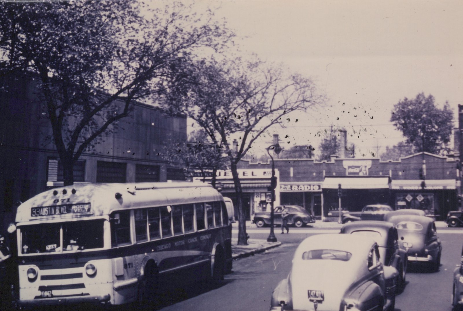 A black and white photo of a vintage bus on a city street, showing its front and side with the number 871 and 'Chicago Motor Coach Company' lettering. Cars from the 1950s and storefronts line the street.