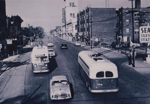 A black-and-white photo showing two trolleybuses on a city street with multiple vintage automobiles and a Sears store in the background.