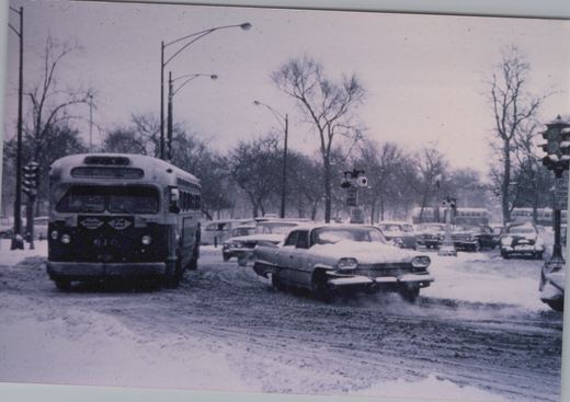 Front view of a vintage city bus driving on a snow-covered street, with classic cars and bare trees in the background.