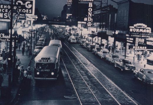 A black and white photo shows a vintage CTA bus labeled '8 HALSTED 79TH ST' and number 638 on a city street at dusk, surrounded by parked cars, pedestrians, and glowing signs for businesses like Sears and Norman Jewelers. The street has embedded trolley tracks, and the scene is bustling with activit