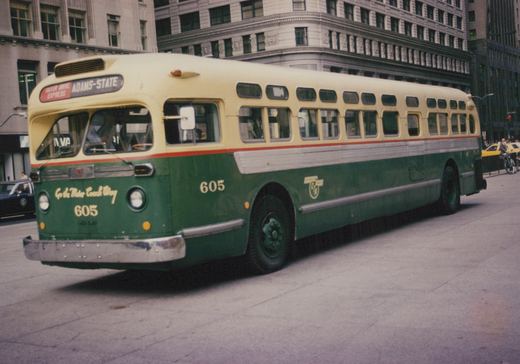 A green and cream Chicago Transit Authority bus, number 605, is shown from a three-quarter front view on a city street with a yellow taxi and a police car in the background.