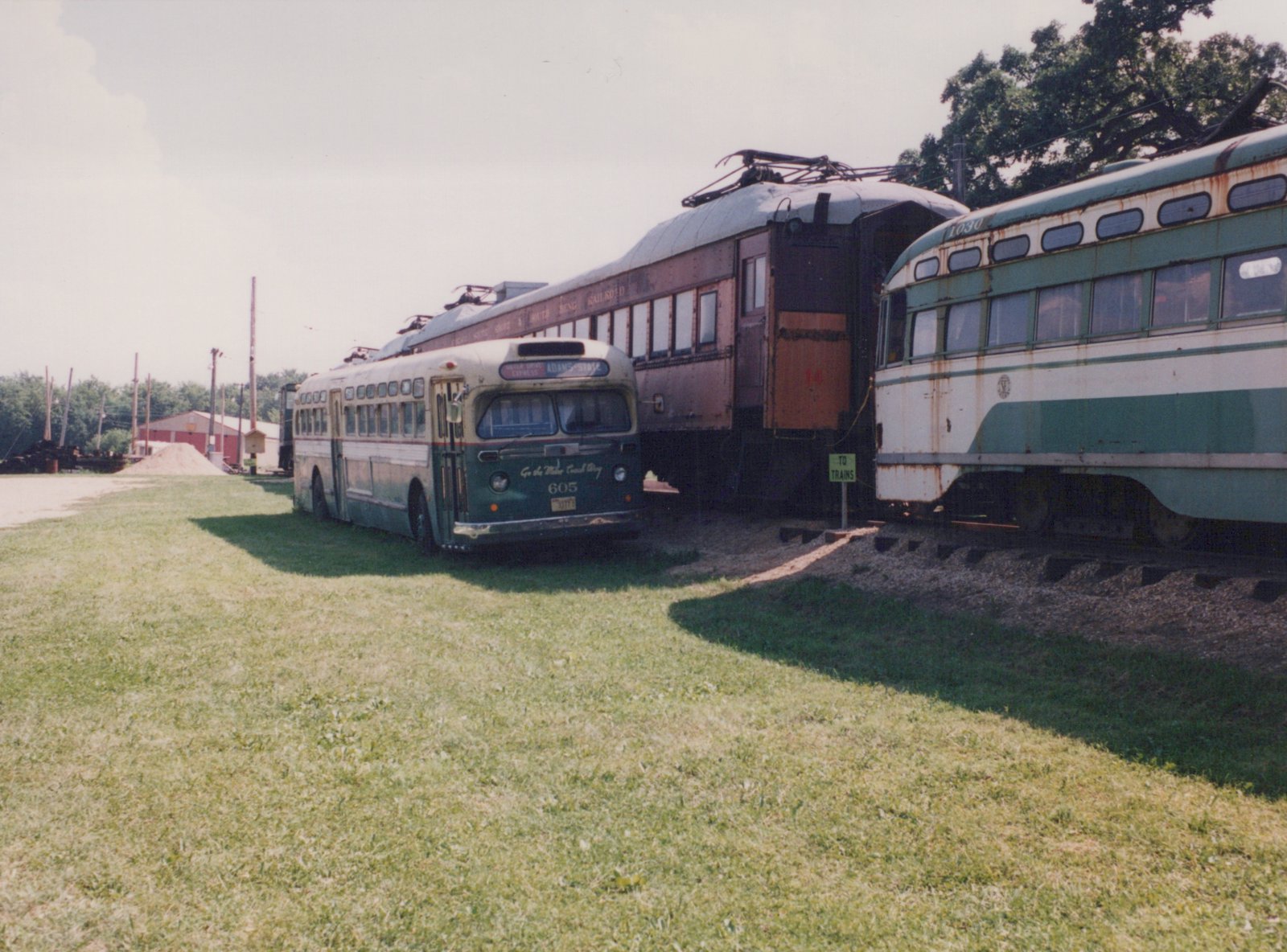 A green and cream bus with the number 605 parked next to two interurban cars with the numbers 1030 and 14 on a sunny day in a grassy yard.