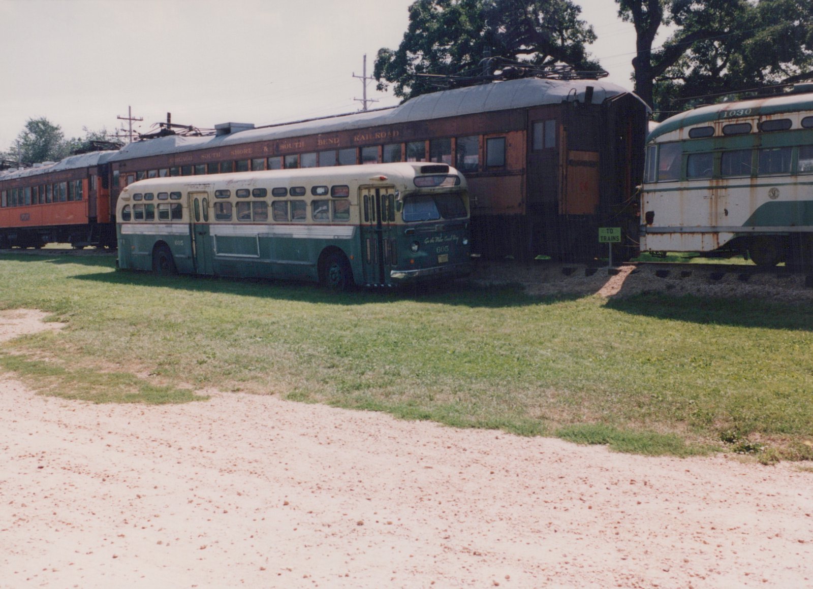A vintage green and cream bus numbered 605 sits on a grassy area beside two old railroad cars, one red and one green and white, with a sign that reads 'TO TRAINS'.