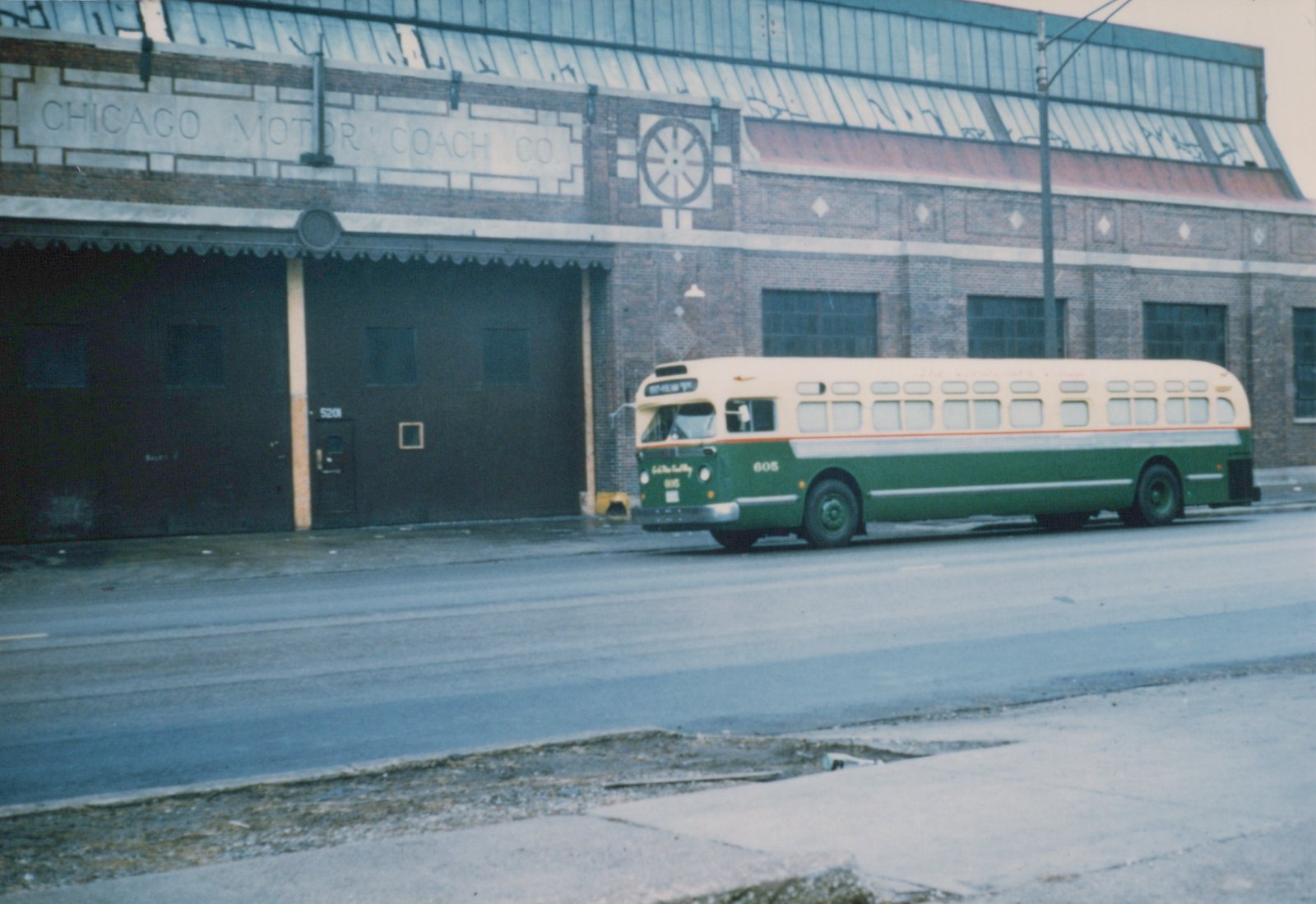 A green and cream Chicago Motor Coach Company bus, number 605, is parked on a wet street in front of a large brick building with 'CHICAGO MOTOR COACH CO.' lettered on its facade.