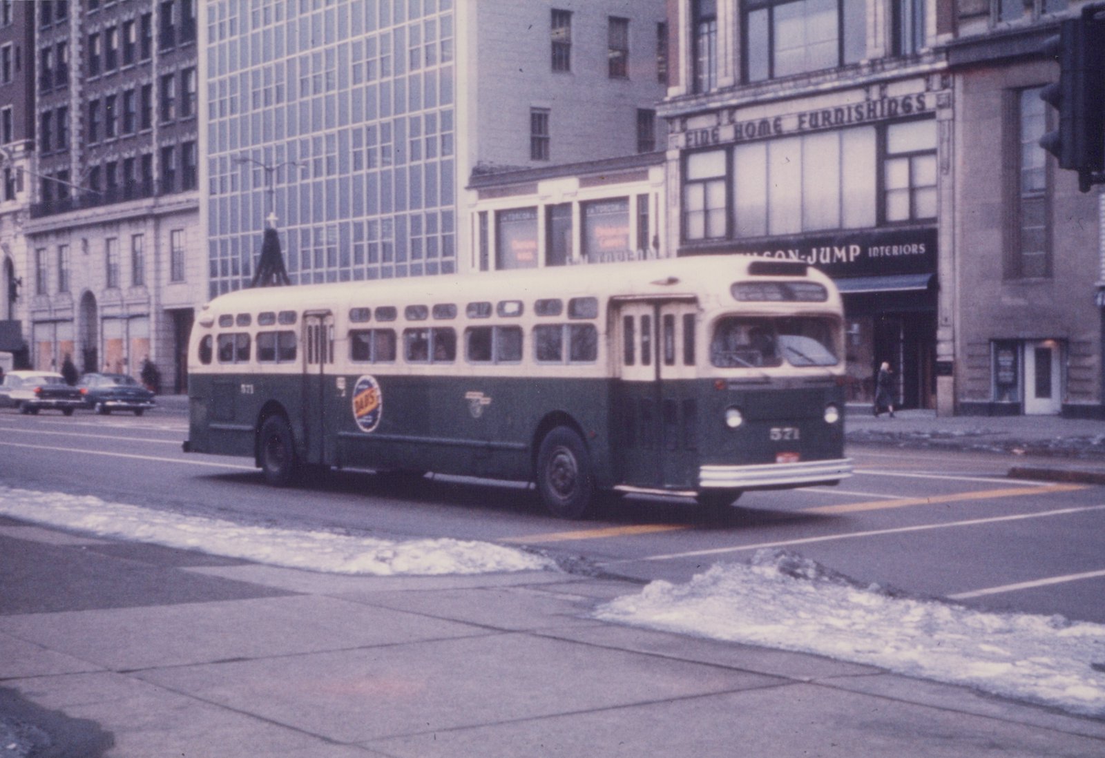 A green and cream motor bus, number 571, drives down a city street with snow on the sidewalks and classic cars parked in the background.