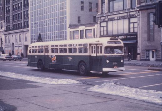 A green and cream motor bus, number 571, drives down a city street with snow on the sidewalks and classic cars parked in the background.