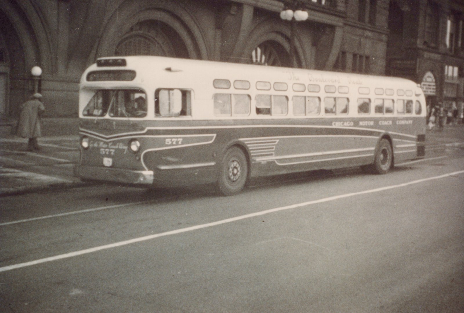 A black and white photo of a vintage Chicago Motor Coach Company bus, number 577, parked on a city street with a person in a coat nearby.