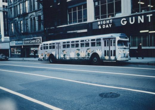 A white bus with a cartoon mural on its side is on a city street, with storefronts and pedestrians in the background.