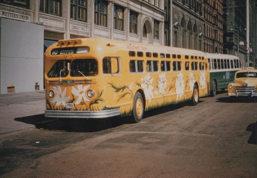 A yellow CTA bus with floral decorations and a 'Make Chicago America's Cleanest City' slogan is parked on a city street, with a green and white bus and a yellow taxi cab visible in the background.