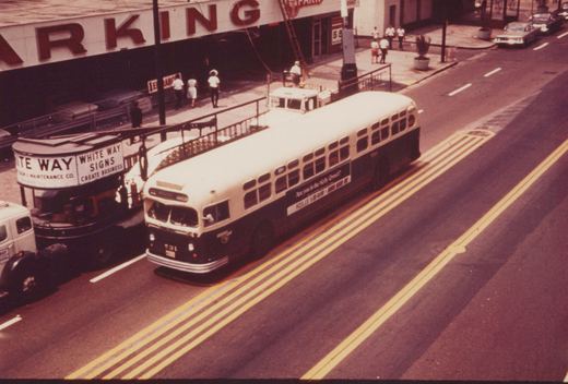 A vintage city bus with the number 31 on its front and side drives down a street lined with parked cars and pedestrians, with a large 'PARKING' sign visible above.