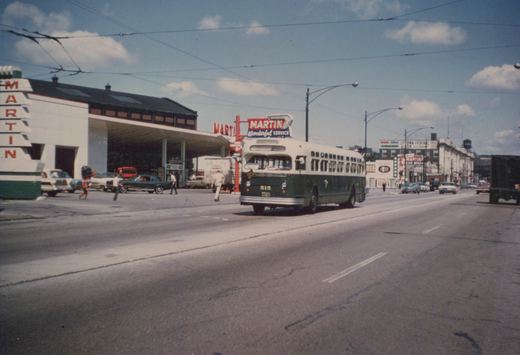 A green and cream Chicago Transit Authority bus numbered 515 travels down a city street on a sunny day, passing a Martin gas station with several 1960s cars and pedestrians nearby.