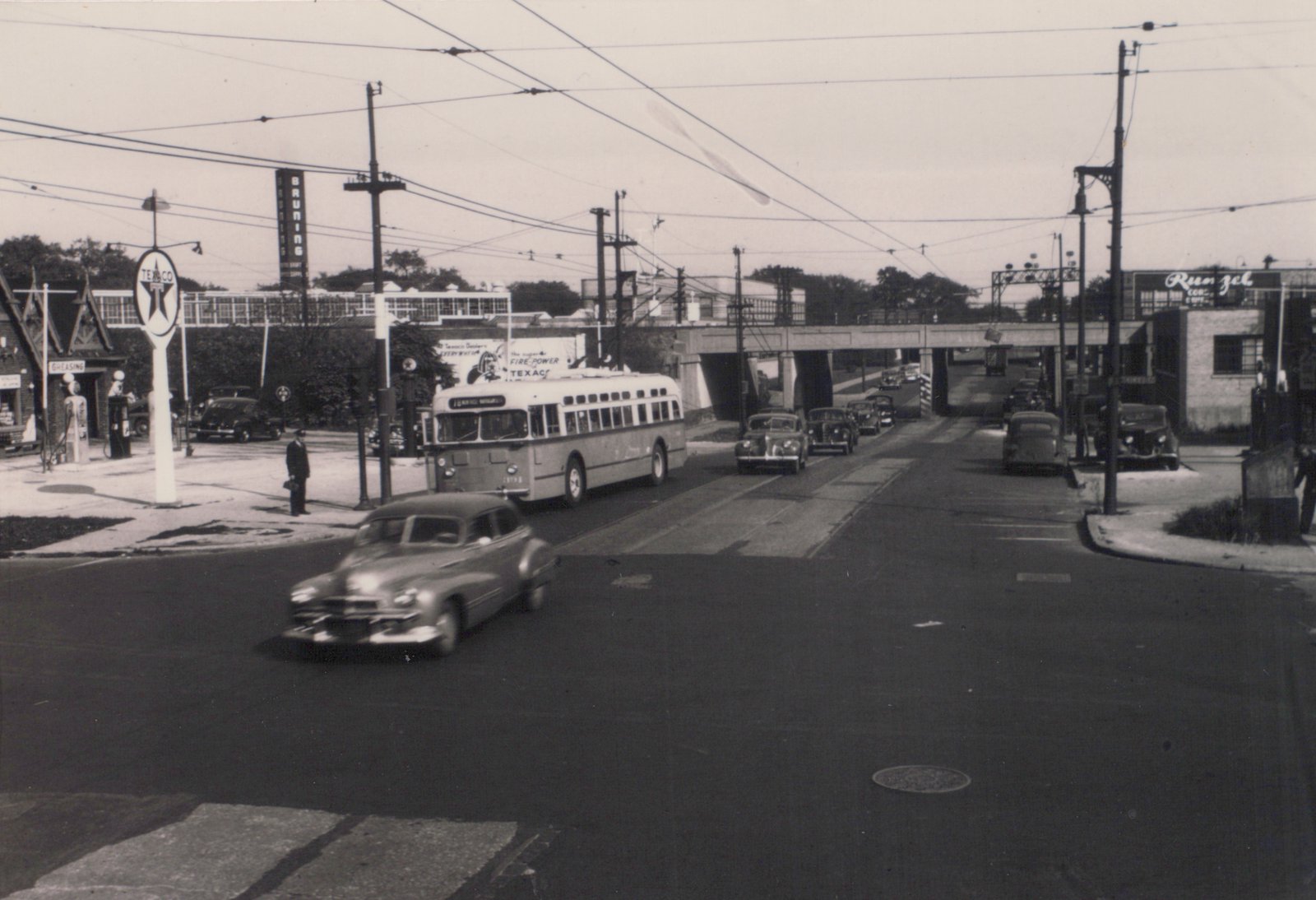 Black and white photo of a CTA bus on a city street with overhead wires, an overpass in the background, and vintage cars.