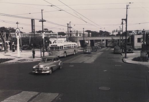 Black and white photo of a CTA bus on a city street with overhead wires, an overpass in the background, and vintage cars.