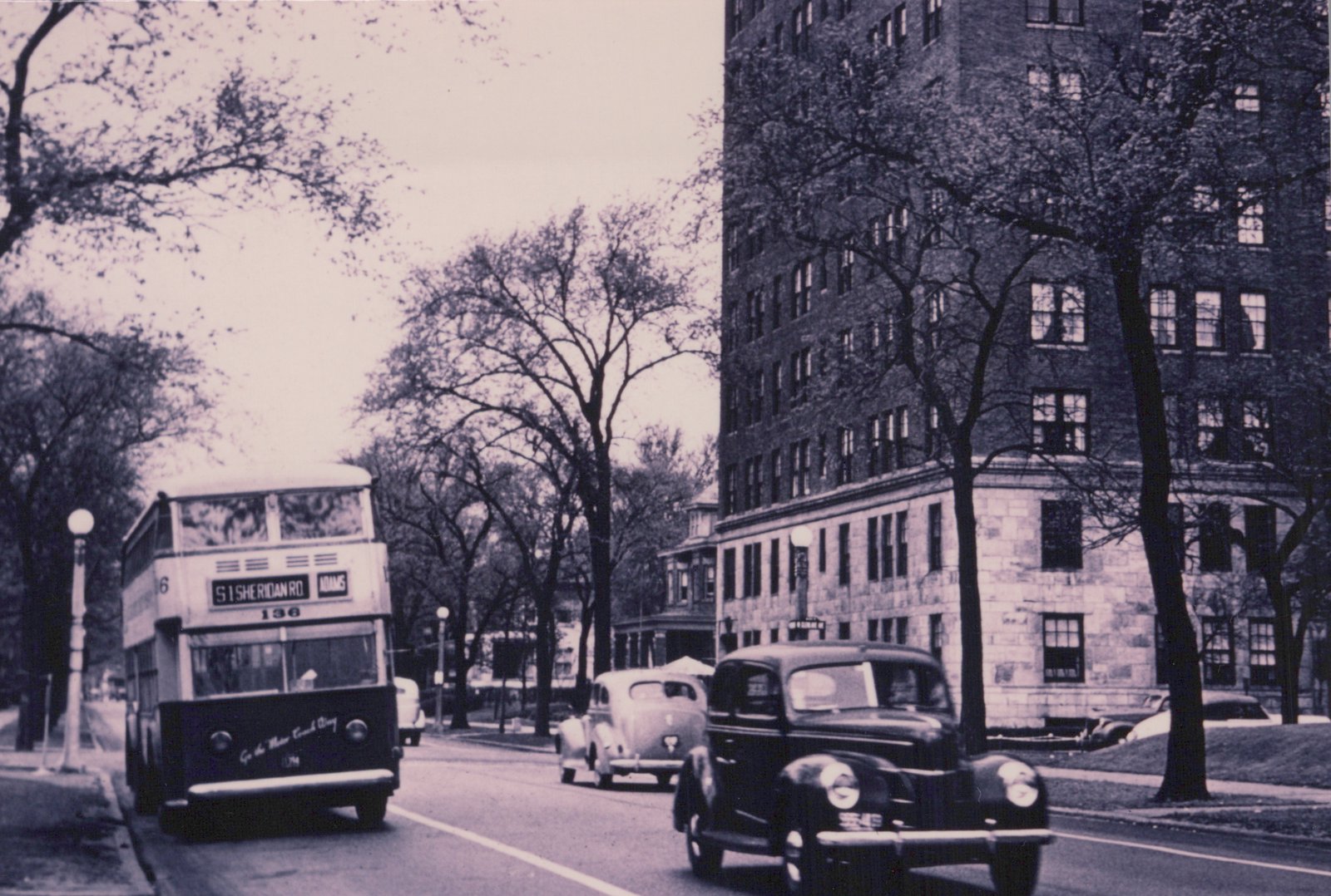 A vintage black-and-white photo shows a double-decker bus on a city street with trees and a large brick building, alongside several 1930s automobiles.