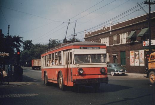 A red and cream trolleybus on a sunny day, with overhead wires, passengers inside, and vintage cars and buildings in the background.