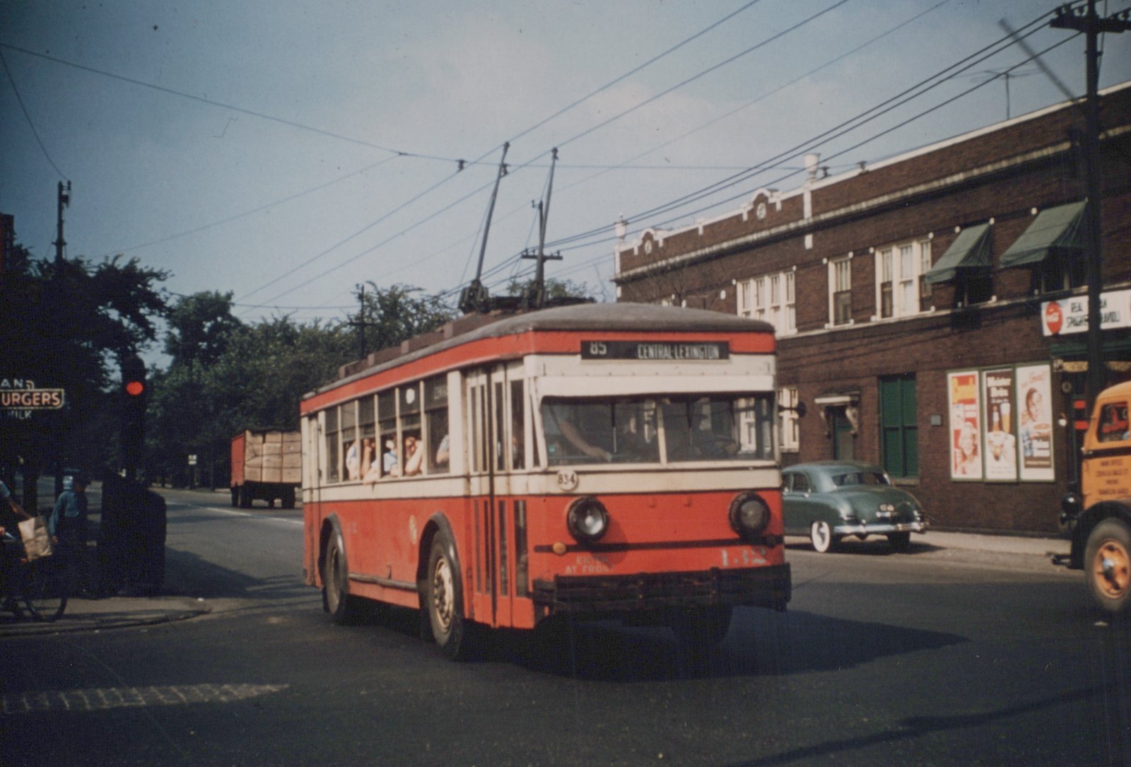 A red and cream trolleybus on a sunny day, with overhead wires, passengers inside, and vintage cars and buildings in the background.