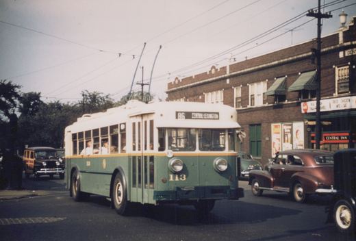 A green and cream CTA trolleybus with the number 113 on the front and route 86 to Central-Lexington displayed, driving down a street lined with brick buildings and vintage cars.