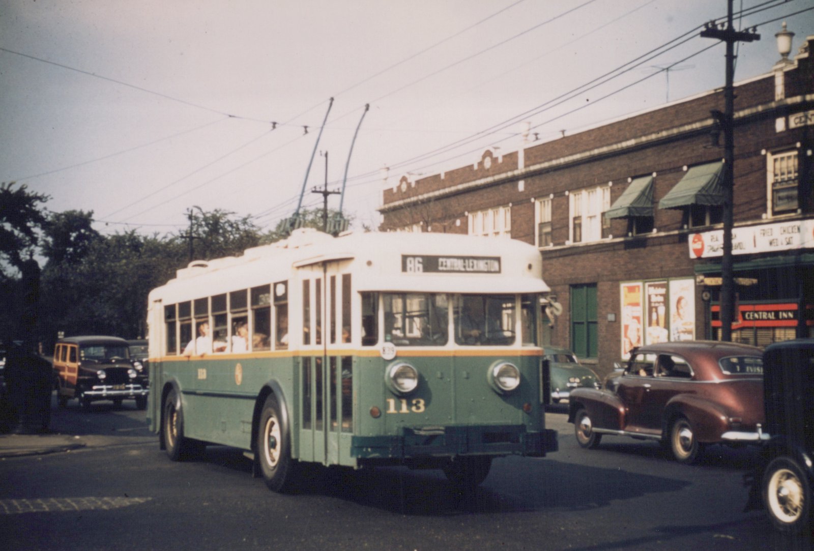 A green and cream CTA trolleybus with the number 113 on the front and route 86 to Central-Lexington displayed, driving down a street lined with brick buildings and vintage cars.