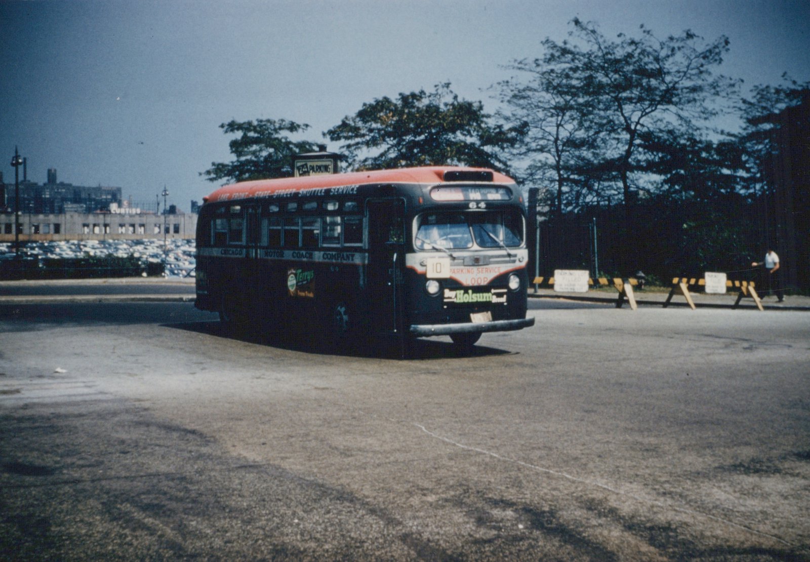 A vintage red-roofed Chicago Motor Coach Company bus, number 64, is parked on a paved lot, with a man walking nearby and trees and buildings in the background.