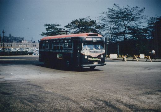A vintage red-roofed Chicago Motor Coach Company bus, number 64, is parked on a paved lot, with a man walking nearby and trees and buildings in the background.