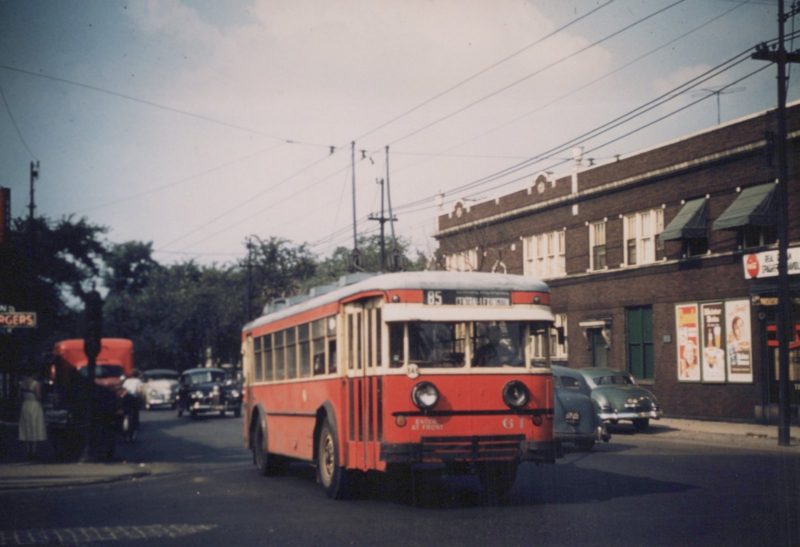A red and cream Chicago Transit Authority trolleybus, number 61, on route 85, is driving on a city street with vintage cars and storefronts, under clear skies.