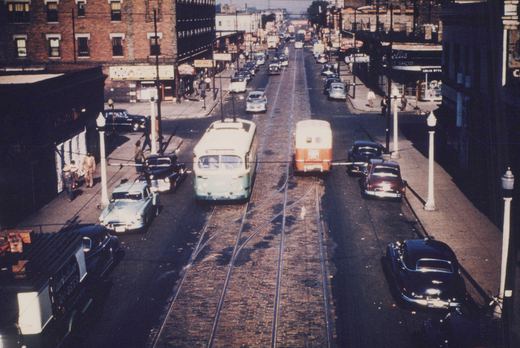 An elevated view of a street in Chicago with two trolleybuses on tracks, surrounded by classic cars and onlookers on the sidewalks.